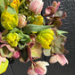 A close-up of a spring flower arrangement featuring bright yellow tulips, pink-and-white hellebores, and green leaves, set against a dark charcoal wood plank background.